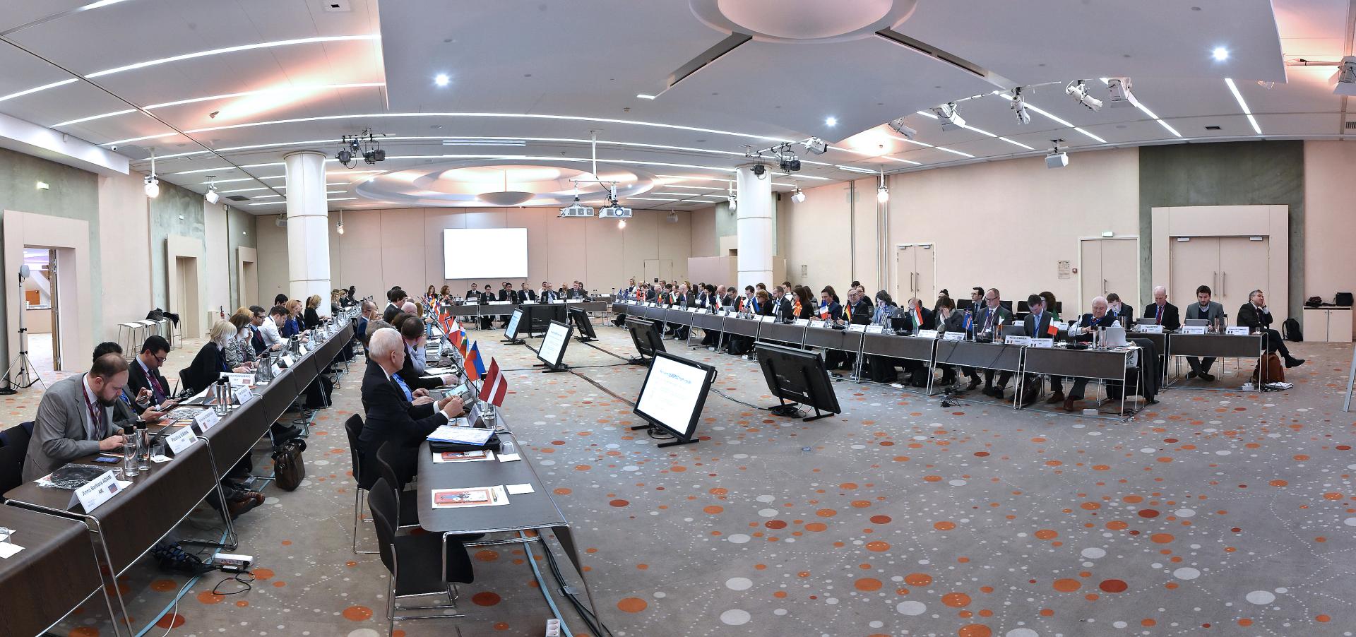 The image shows a meeting hall with multiple participants sitting at a U-shaped table group