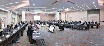 The image shows a meeting hall with multiple participants sitting at a U-shaped table group