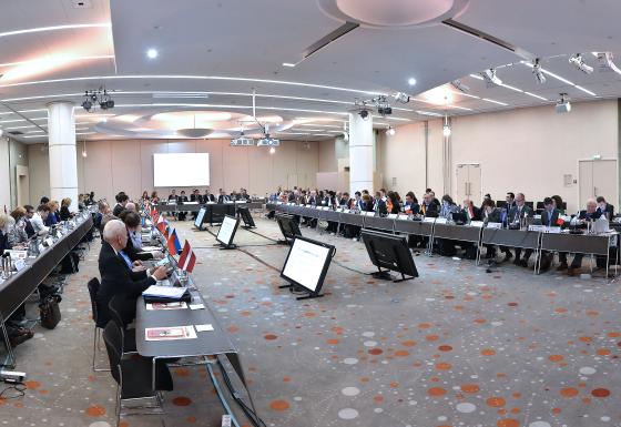 The image shows a meeting hall with multiple participants sitting at a U-shaped table group